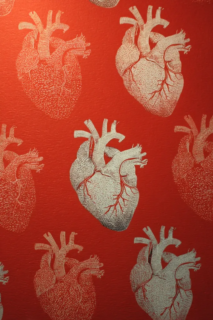 A close-up shot reveals a vibrant red surface adorned with a repeating pattern of anatomically accurate hearts.  The hearts are rendered in a light off-white or silver, creating a striking contrast against the deep red background. The detailed linework emphasizes the heart's chambers and vessels, suggesting a medical or anatomical theme. The overall effect is bold and slightly macabre, possibly suitable for wallpaper or decorative fabric.