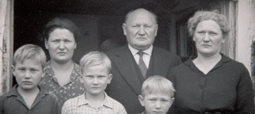 A black and white family portrait shows a stern-faced older couple flanked by their children and grandchildren. The older man is wearing a suit and tie. The older woman wears a dark blouse. The younger generation are dressed in more casual attire. The image suggests a formal or commemorative photograph. The expressions are serious, hinting at a possibly somber occasion. The overall tone is one of quiet dignity and family unity.