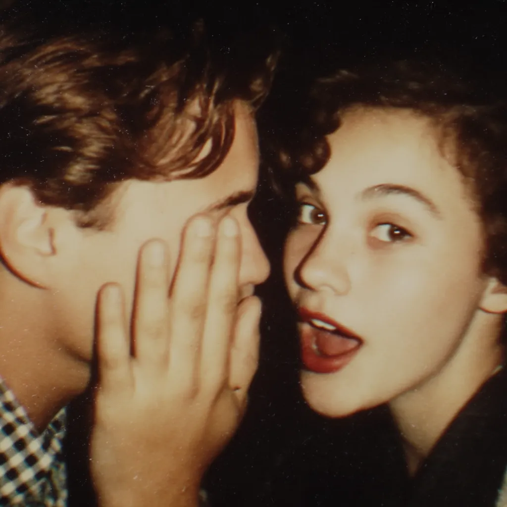 A close-up shot captures a young couple, their faces nearly touching. The woman, with dark, curly hair, leans in, her expression one of excited anticipation.  The man, with light brown hair, has his hand gently covering part of his face as she whispers a secret into his ear. The image's vintage quality and warm tones enhance the intimate and nostalgic mood.