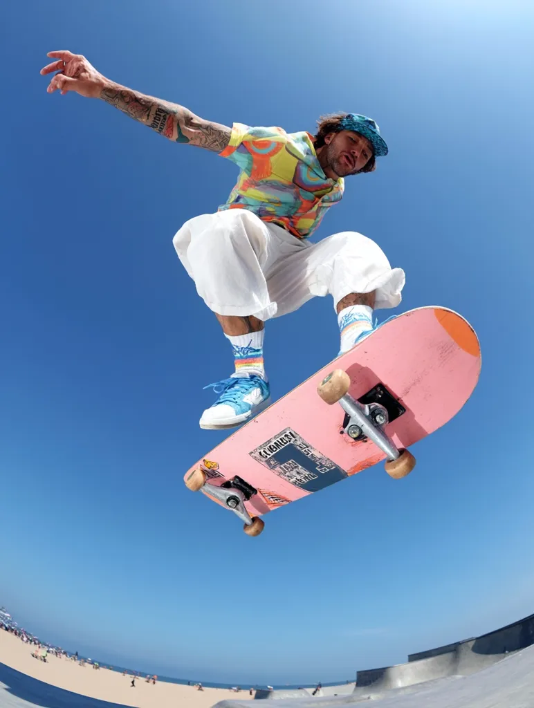 A skateboarder, wearing a vibrant patterned shirt and white pants, executes a stylish aerial maneuver over a skate park.  The bright pink skateboard is in sharp focus against the clear blue sky.  A beach is visible in the background, adding to the sunny, energetic atmosphere of the image.  The skater's tattoos are partially visible, contributing to a sense of individuality and freedom.