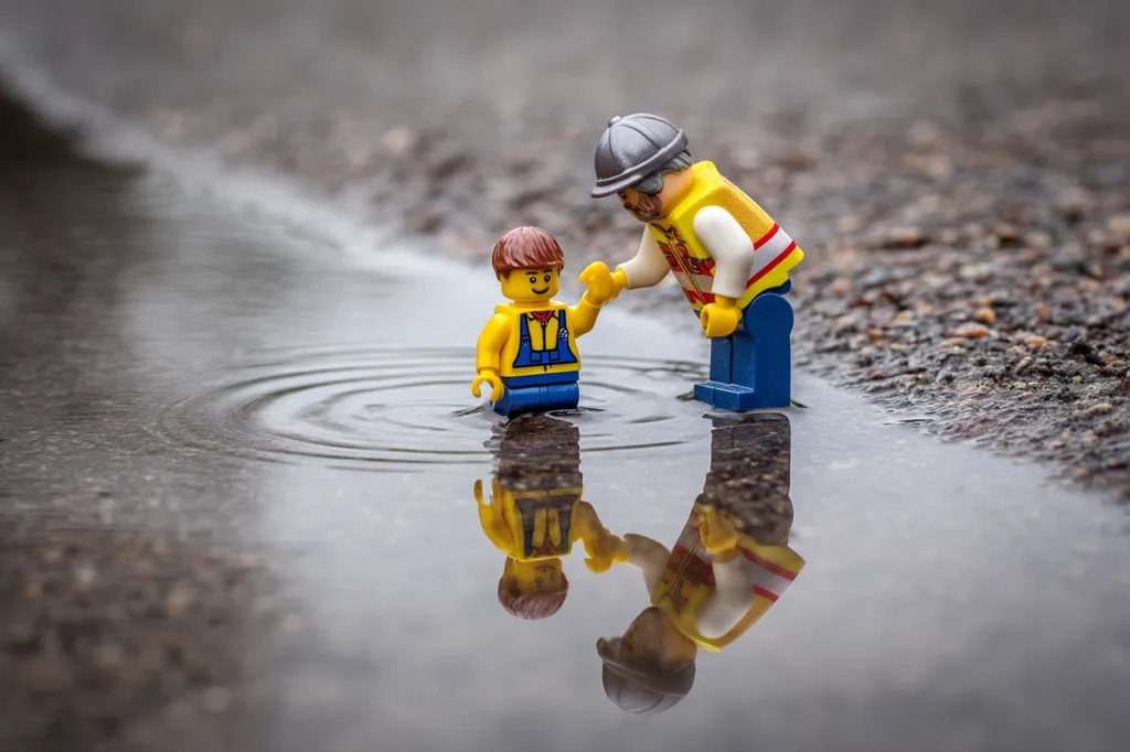 Two Lego figures, one adult and one child, stand in a puddle.  The adult, wearing a hard hat and safety vest, reaches out to help the smaller figure.  Their reflections are visible in the water, creating a mirrored image.  The scene is set outdoors on a wet, dark street.  The overall mood is gentle and heartwarming.