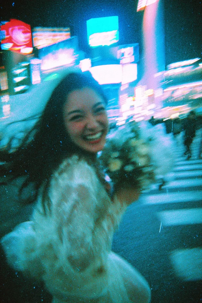 A blurry, vibrant photo captures a young woman smiling brightly, holding a bouquet of flowers.  She wears a light, lacy dress. The background is a bustling city street at night, with neon lights creating a hazy, motion-blurred effect. The overall mood is joyful and energetic, with a dreamy, nostalgic quality due to the film grain and motion blur.