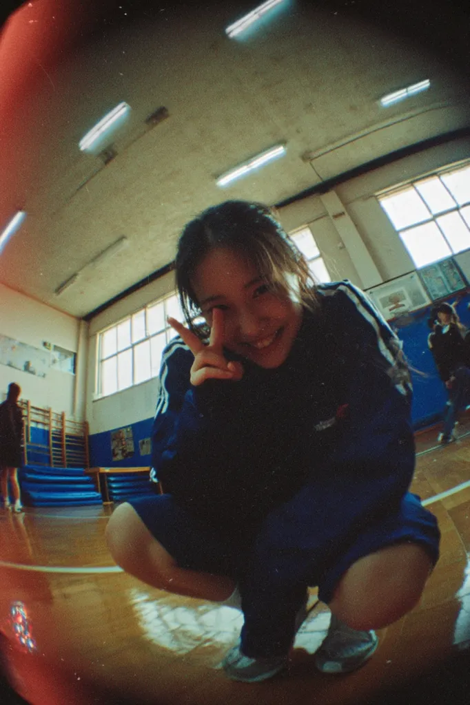 A young woman crouches in a gymnasium, giving a peace sign to the camera.  She's wearing a dark blue tracksuit with white stripes. The gym is brightly lit with fluorescent lights, and the walls are a muted beige.  The photo is taken with a fisheye lens, creating a distorted perspective. Another person is visible in the background. The overall mood is casual and playful.