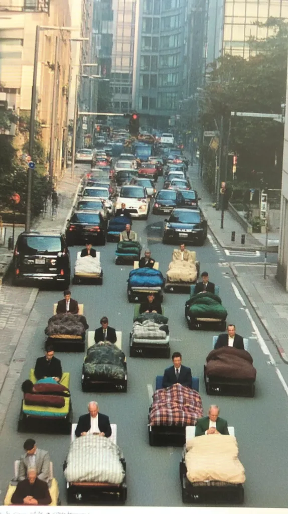 A city street scene shows numerous small vehicles, each carrying a person bundled in blankets on what appears to be a makeshift bed.  The vehicles are moving slowly through congested traffic amongst larger cars.  The setting suggests a Japanese city, with tall buildings and a somewhat dated aesthetic. The image conveys a surreal and humorous juxtaposition of everyday commuting with unusual comfort measures.