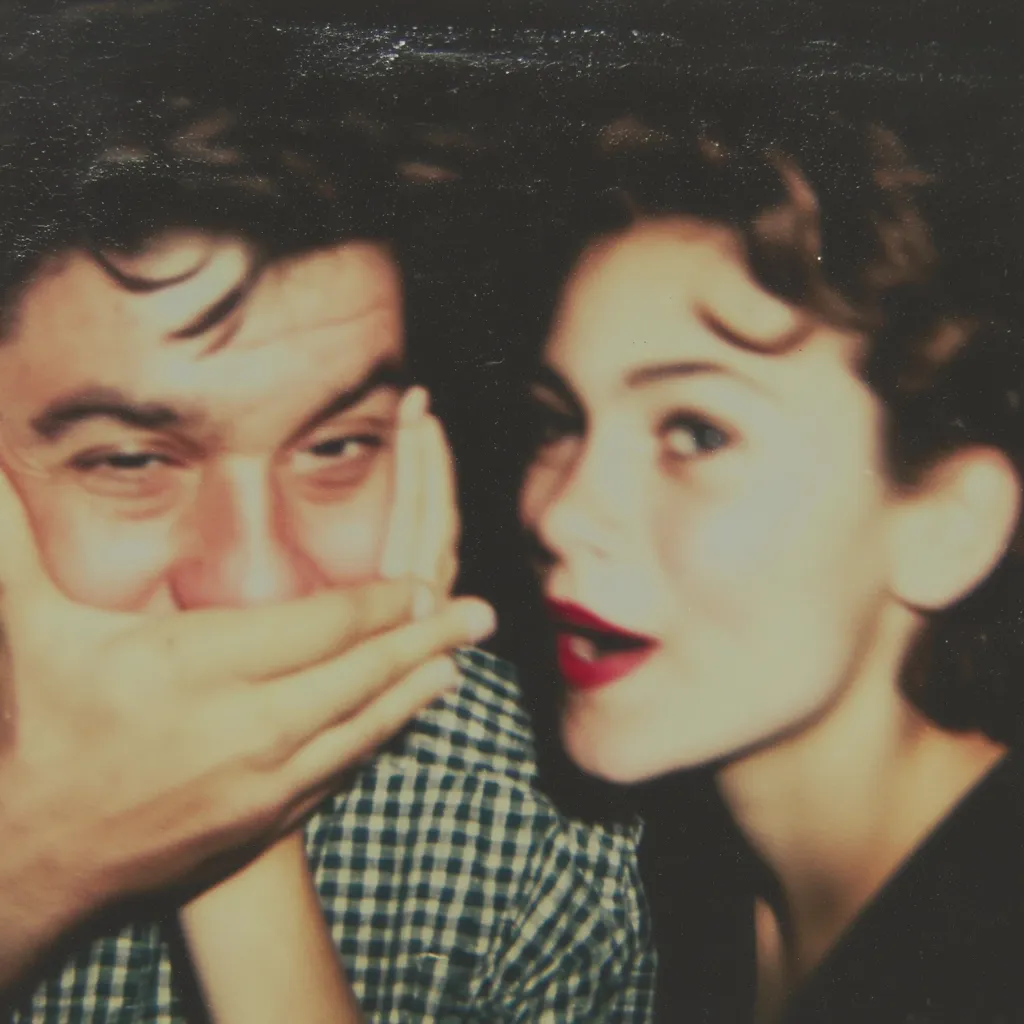 A close-up, slightly blurry vintage photograph depicts a young couple. The man, wearing a plaid shirt, playfully covers his mouth with his hands.  The woman, with dark, wavy hair and red lipstick, looks directly at the camera, possibly whispering a secret.  The image has a faded, nostalgic quality, characteristic of old photographs.