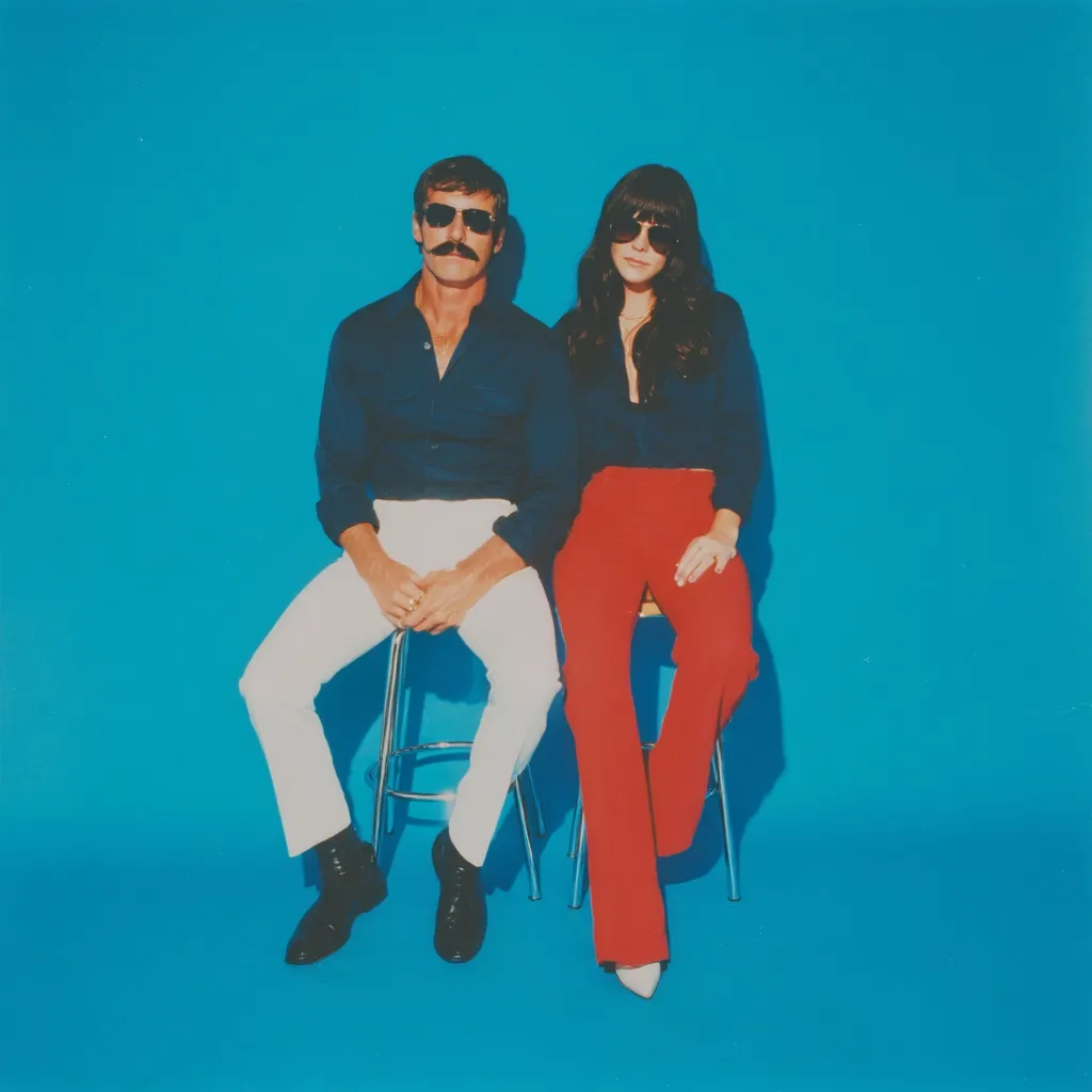A man and woman sit on separate stools against a bright blue background.  The man, with a dark mustache and sunglasses, wears a dark blue shirt and white pants. The woman, with long dark hair and sunglasses, wears a dark blue shirt and bright red pants. Their 1970s-style attire and pose create a retro aesthetic.