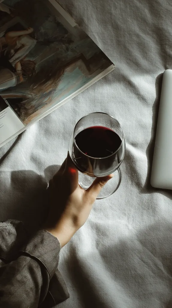 A hand holds a glass of red wine on a bed.  A partially visible book with a painting lies nearby, along with a laptop. The scene is bathed in soft, natural light, casting shadows on the light-grey linen sheets. The overall aesthetic is calm and relaxed, suggesting a moment of quiet contemplation or leisure.