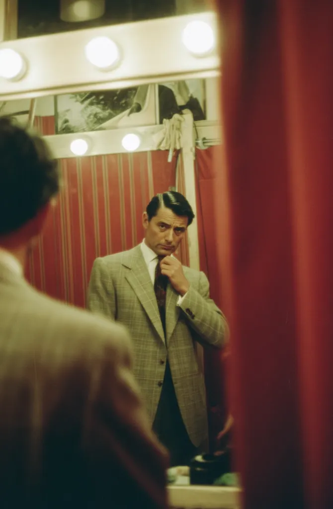 A man in a light gray suit adjusts his tie while looking at his reflection in a mirror. The mirror is surrounded by bright lights, and the background features red-striped walls and curtains. The man's expression is thoughtful, and his gaze is directed towards the viewer, creating a sense of intimacy and introspection.  The image evokes a feeling of backstage preparation or a moment of private reflection.