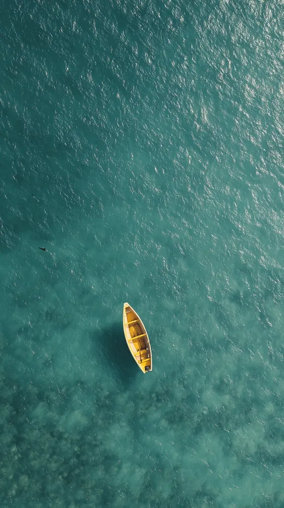 An aerial view captures a solitary yellow rowboat adrift in a tranquil expanse of teal water. The water's surface displays subtle textural variations, suggesting gentle currents.  The boat is positioned centrally, creating a sense of serenity and isolation. The overall image evokes a feeling of peace and calm.