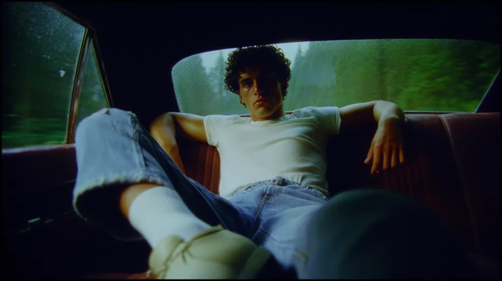 A young man with curly hair relaxes in the backseat of a car. He wears a white t-shirt and jeans, his legs stretched out. The car's interior is dark, contrasting with the vibrant green landscape visible through the rain-streaked window.  His expression is serious, almost pensive, as he gazes forward. The overall mood is one of quiet contemplation and a sense of journey.