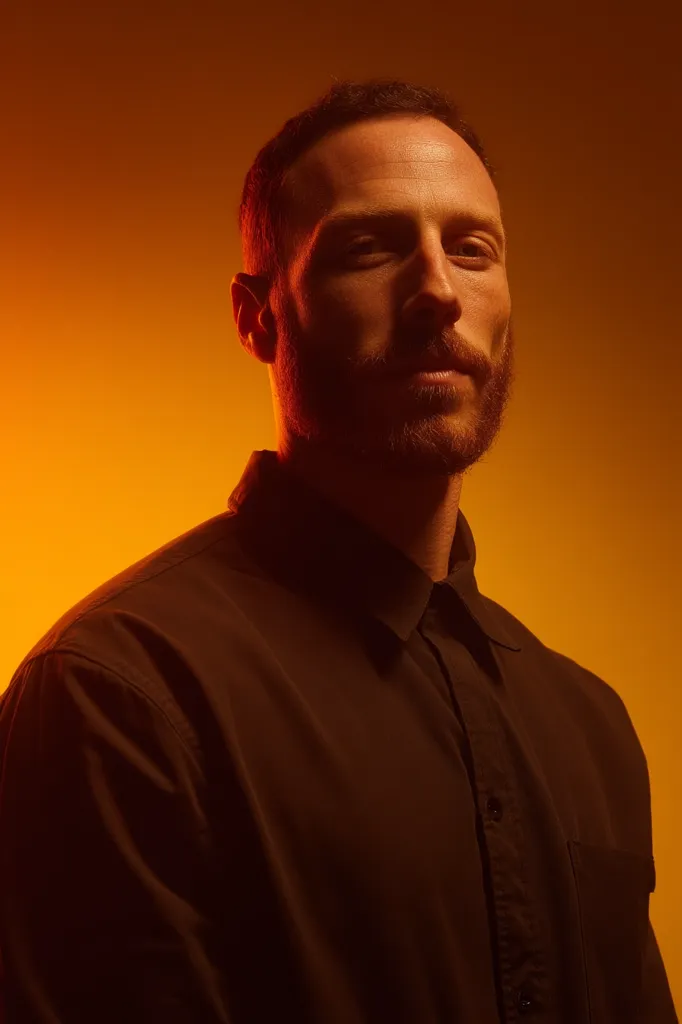 A man with a beard and short dark hair is shown from the chest up against an orange background. He is wearing a dark collared shirt.  The lighting accentuates his facial features and creates a dramatic, moody portrait. His expression is serious and his gaze is directed slightly to his right.