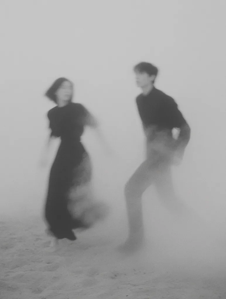 A blurry black and white photo depicts a man and woman walking on a beach, enveloped in a misty fog.  Their figures are indistinct, creating a sense of mystery and ethereal beauty.  The couple's dark clothing blends with the hazy background, enhancing the dreamlike atmosphere.  The overall impression is one of romance and fleeting moments.