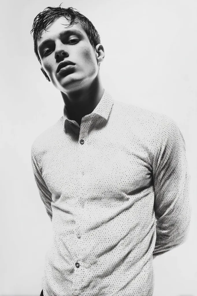 A black and white, close-up portrait of a young man. He has short, wet hair and is wearing a light-colored, button-down shirt with a subtle pattern. His expression is serious, and his gaze is directed slightly downward and away from the camera. The image has a moody and stylish feel, emphasizing the man's features and the texture of his shirt.  The background is plain and unblemished.