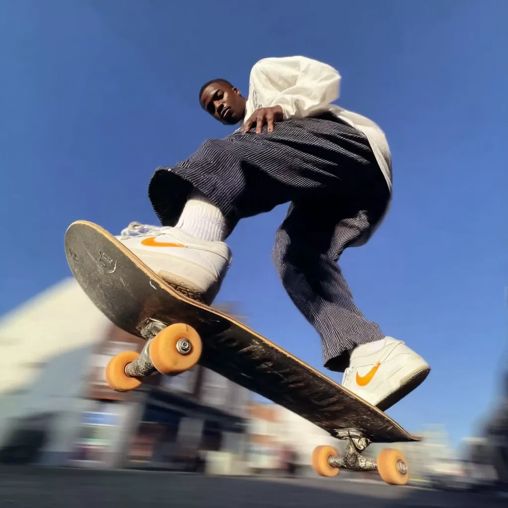 A low-angle shot captures a young, dark-skinned man skateboarding.  He's dressed in loose-fitting, striped pants, a cream-colored shirt, and white Nike sneakers with orange accents.  The skateboard is in motion, with the background blurred, suggesting speed. The bright sun and clear blue sky provide a vibrant backdrop.  The overall image is dynamic and energetic.