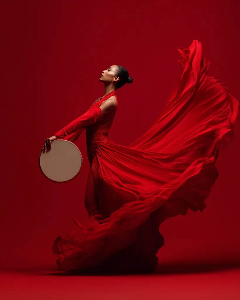 A woman in a flowing red gown holds a tambourine.  The dress billows dramatically around her as she stands against a crimson backdrop.  Her pose is elegant and graceful, suggesting movement and artistry. The overall image is one of passion and vibrant energy. The red color dominates, creating a powerful and visually striking effect.