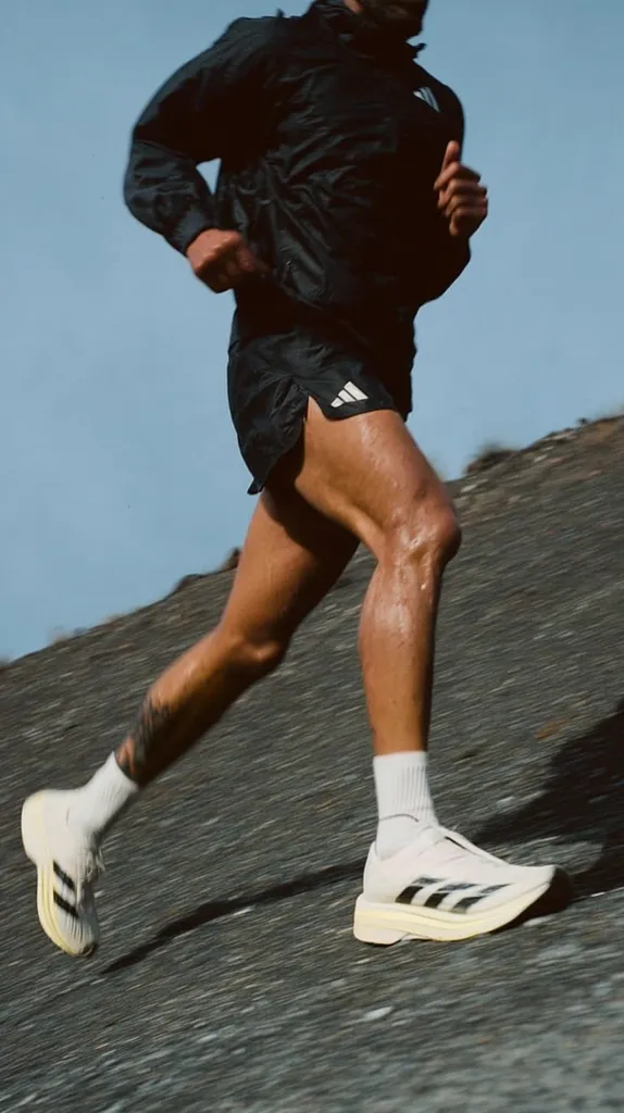 A runner, wearing a black Adidas jacket and shorts, strides along a dark road.  Sunlight glints off his sweat-dampened legs. He's wearing white Adidas running shoes with black stripes. The scene is dynamic, capturing the motion of his run against a backdrop of a clear blue sky and a gently sloping hill.  The overall mood is one of athleticism and freedom.