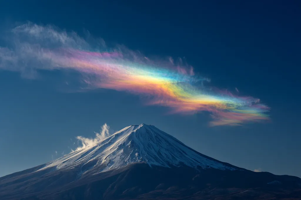 A breathtaking view of Mount Fuji, its snow-capped peak rising majestically against a vibrant sky.  A stunning iridescent cloud, displaying a spectrum of pastel colors, arcs gracefully above the mountain, creating a surreal and awe-inspiring scene. The contrast between the dark mountain and the brightly colored cloud is striking and unforgettable.
