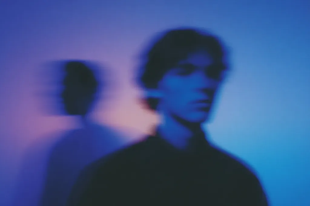 The image is a blurry portrait of a person, possibly a man, bathed in shades of blue and purple light.  The subject is slightly out of focus, creating a dreamlike or ethereal effect.  A second, even more indistinct figure is visible in the background, adding to the mysterious atmosphere. The overall mood is somber and introspective.