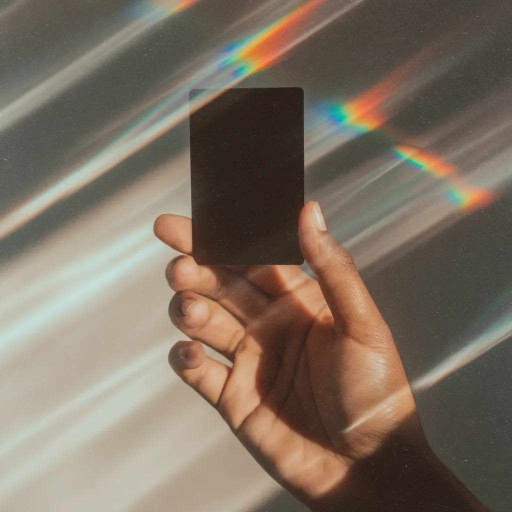 A dark rectangular object, possibly a card or phone, is held in a person's hand.  The hand is dark-skinned and the background features streaks of vibrant, rainbow-like light, creating a dreamy, ethereal effect. The image is stylistic and atmospheric, with a focus on the contrast between the dark object and the bright, colorful background.