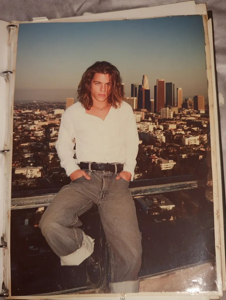 A young man with long brown hair sits on a railing, hands in pockets, overlooking the Los Angeles cityscape.  He wears a white button-down shirt and loose-fitting jeans.  The city skyline, including several skyscrapers, stretches behind him under a clear, bright sky.  The photo is a vintage print, likely taken from a personal album.