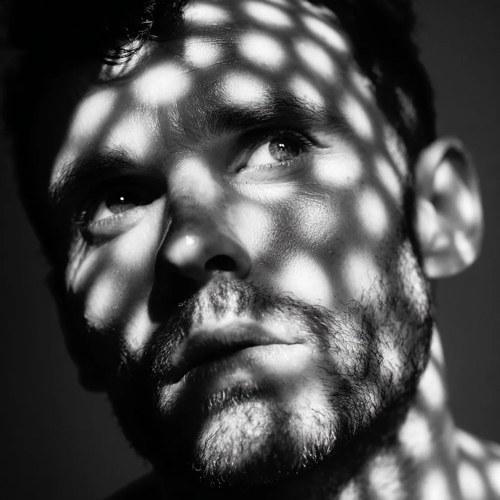 A black and white close-up portrait of a man's face, partially obscured by a patterned shadow cast from a light source.  His expression is contemplative, his gaze slightly upward. The shadows create a dramatic and textured effect on his skin and beard, highlighting the contours of his face. The overall mood is introspective and moody.