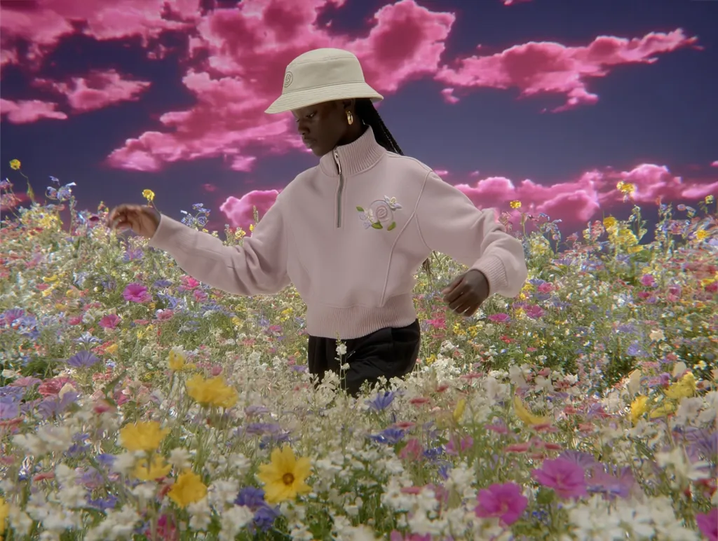 A young woman in a pale pink sweater and bucket hat walks through a vibrant, surreal field of wildflowers.  The sky is a dramatic pink and purple.  She appears to be enjoying the whimsical, dreamlike setting. The overall mood is one of gentle joy and fantasy.