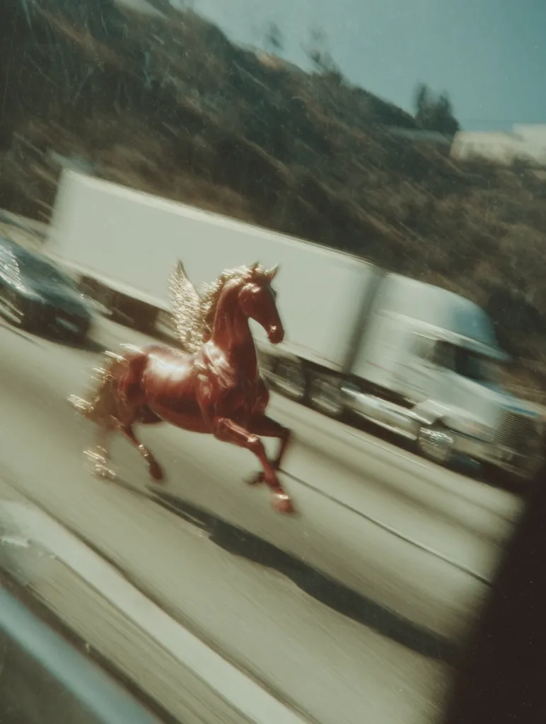 A blurry, motion-filled photograph captures a red pegasus figurine seemingly running alongside a highway.  The background is a fast-paced blur of vehicles, including a large white truck, against a hillside. The image suggests speed and a surreal juxtaposition of the mythical creature and the mundane reality of highway traffic.  The overall tone is dreamlike and slightly nostalgic.