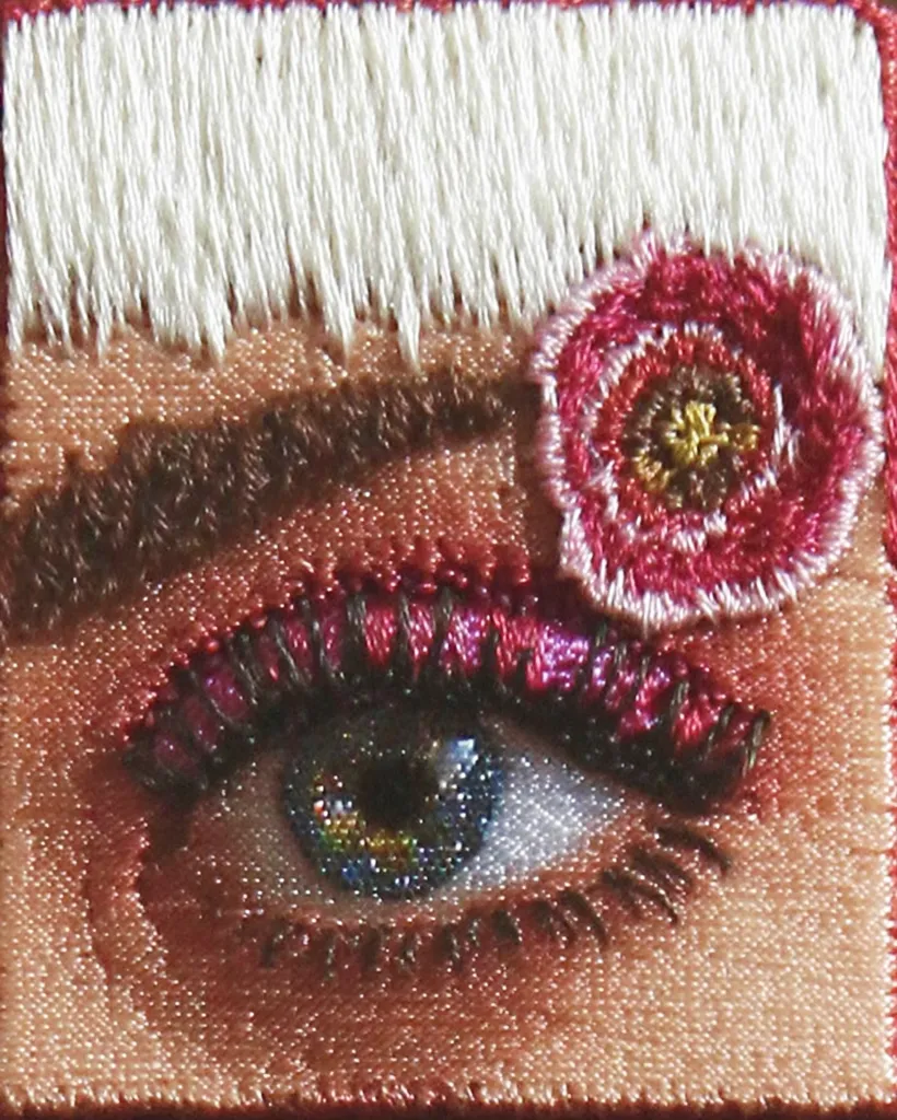 A detailed close-up of an embroidered artwork depicts a woman's eye and brow.  The piece is meticulously crafted using various embroidery stitches to create texture and depth, with a pink flower accent near the temple.  The eye is realistically rendered, showcasing fine details such as individual lashes and a shimmering iris. The overall effect is a miniature portrait, rich in color and texture.