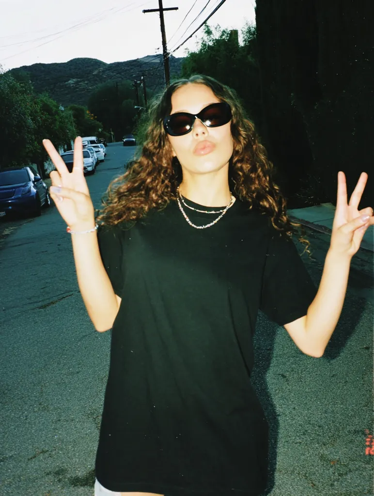 A young woman with long, curly brown hair wears black sunglasses and a loose black t-shirt.  She's making peace signs with both hands. The photo, with a slightly grainy, film-like quality, shows her on a residential street, with cars parked along the curb and hills visible in the background.  She wears delicate necklaces.