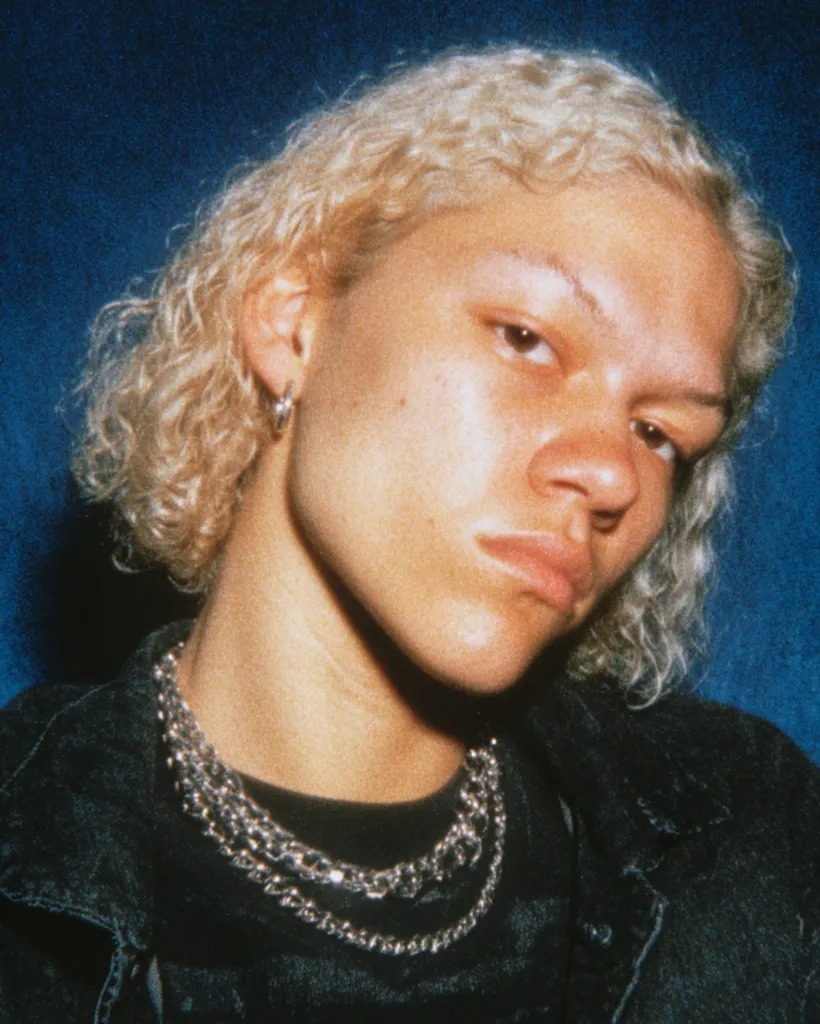 Close-up portrait of a young person with blonde, curly hair. They are wearing a dark denim jacket and a layered silver chain necklace.  Their expression is serious, almost brooding. The image has a slightly grainy, vintage quality, possibly from a film photograph.  A small silver hoop earring is visible in their left ear. The background is a deep blue.