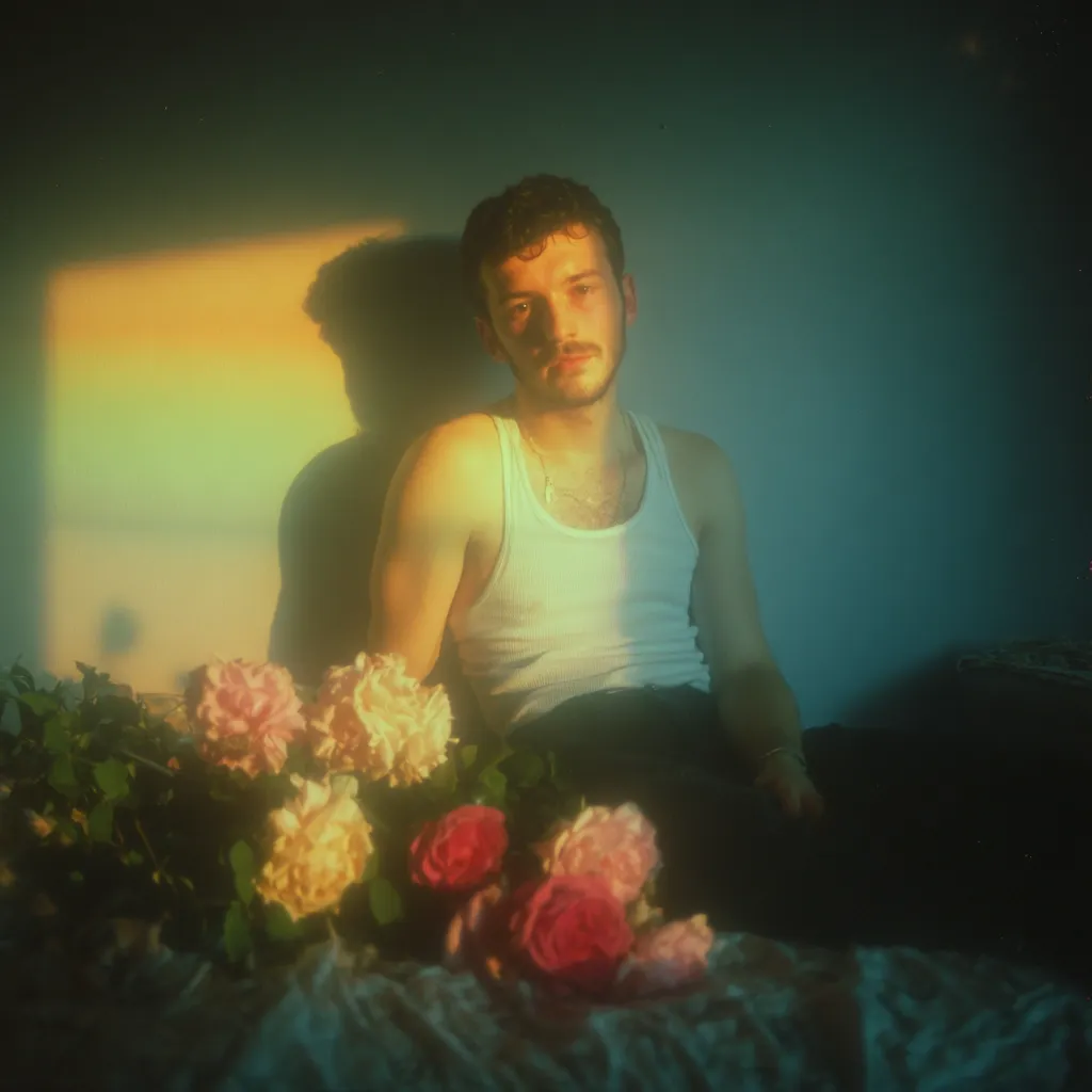 A young man with short brown hair sits on a bed, illuminated by warm, sunlit hues. He's wearing a white tank top and dark pants, a simple silver necklace visible.  A bouquet of pink, white, and rose-colored roses rests on the bed in front of him, adding to the soft, romantic ambiance of the photograph. The walls cast a subtle, rainbow-like reflection on the wall behind him.