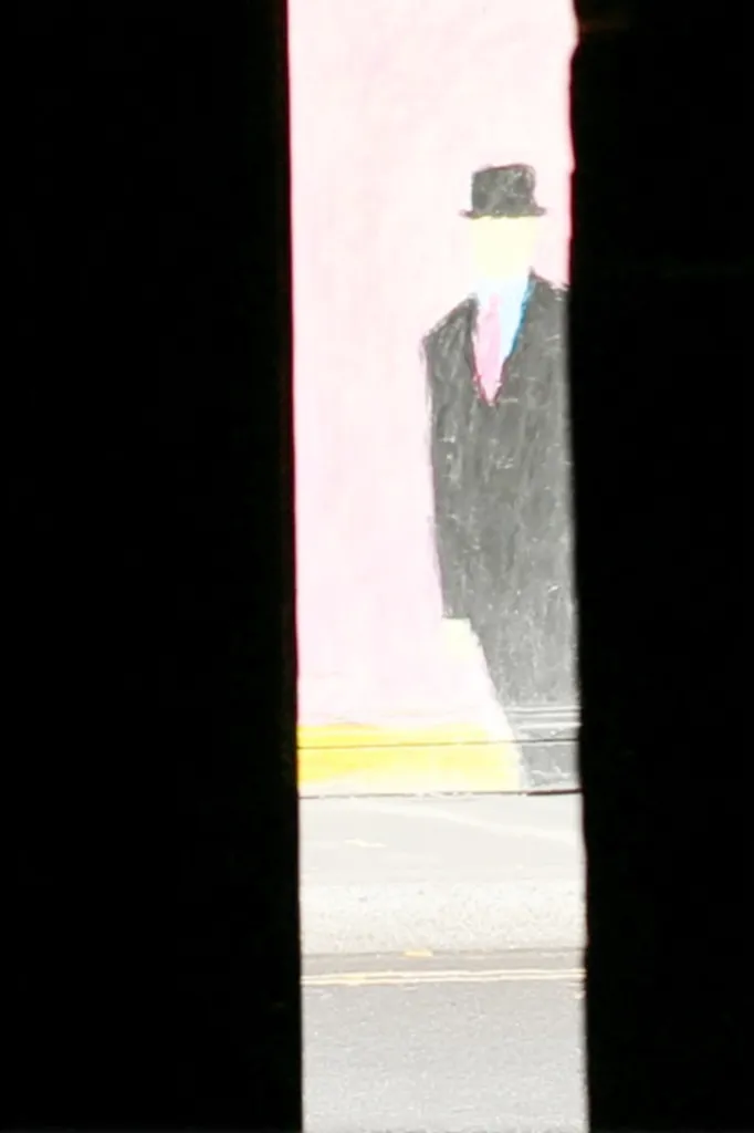 A partially obscured view of a street art painting.  The artwork depicts a simple figure, a man in a suit and bowler hat, standing against a pink background.  The image is framed by a dark, vertical element, possibly a building's edge, which cuts across the view, obscuring parts of the painting and the street beyond. The style is minimalistic, almost childlike in its simplicity.