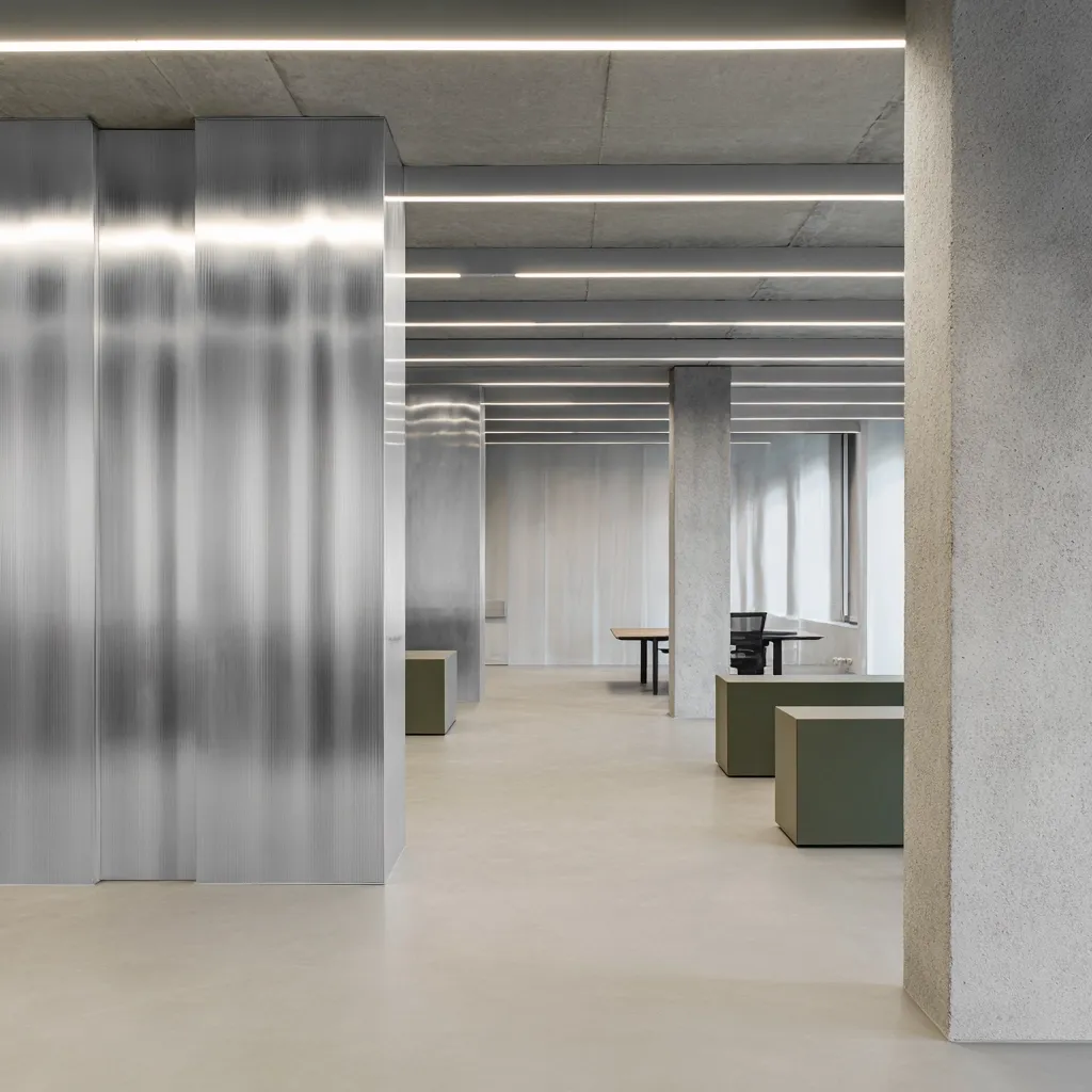 A minimalist office space features a polished concrete floor and ceiling with linear fluorescent lighting.  Large, translucent ribbed panels create partial room dividers.  Concrete columns support the structure.  Simple, olive-green seating elements are scattered throughout the largely empty space, creating a calm and modern atmosphere.  A single dark desk is visible in the background.