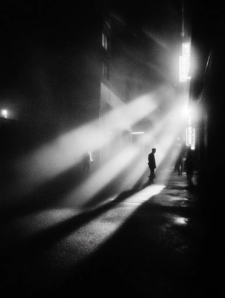A solitary figure stands silhouetted on a foggy city street at night.  Bright beams of light, possibly from street lamps, pierce the mist, creating dramatic streaks of illumination across the dark pavement.  The atmosphere is moody and evocative, with the lone individual appearing small and isolated within the vast, shadowy urban space. Buildings loom in the background, partially obscured by the dense fog.