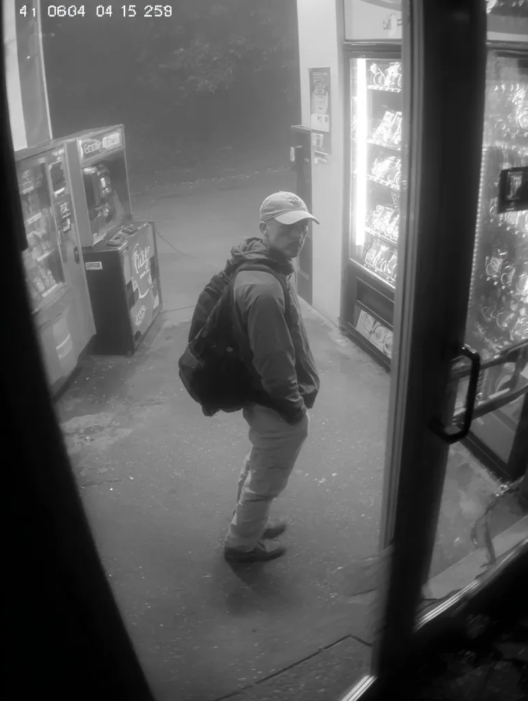 A grainy, black and white security camera image shows a man standing outside a convenience store at night. He wears a baseball cap, a jacket, and light-colored pants. A backpack is slung over his shoulders.  Vending machines are visible inside the store. The image is poorly lit, and the man's features are somewhat obscured.  The timestamp in the corner indicates the time of the image.