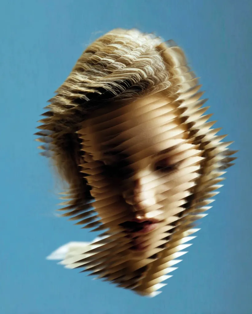 A stylized portrait of a woman's face is presented against a solid blue background.  The image uses a layered, fragmented effect, creating a sense of depth and distortion.  The woman's blonde hair is visible, partially obscuring her features which are subtly visible through the layered effect.  The overall impression is one of abstraction and mystery.
