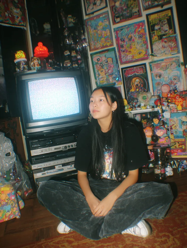 A young woman with long dark hair sits cross-legged on a floor in front of a vintage television displaying static.  She wears a black t-shirt and dark corduroy pants. The background is a vibrant wall covered in numerous framed anime and cartoon posters and collectibles, creating a cluttered yet nostalgic atmosphere.  The room is dimly lit, highlighting the eclectic collection of memorabilia.