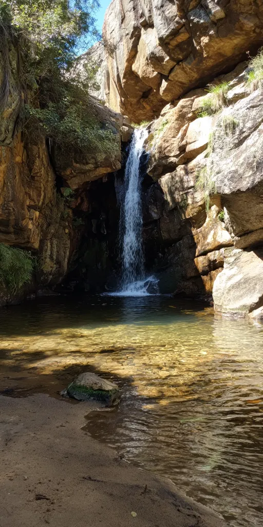 A serene waterfall cascades down rocky cliffs into a clear, shallow pool.  Sunlight illuminates the golden-hued water, creating a tranquil atmosphere.  Lush green vegetation clings to the rocky crevices, contrasting with the dry, sandy bank at the bottom. The overall scene is one of natural beauty and peaceful seclusion.