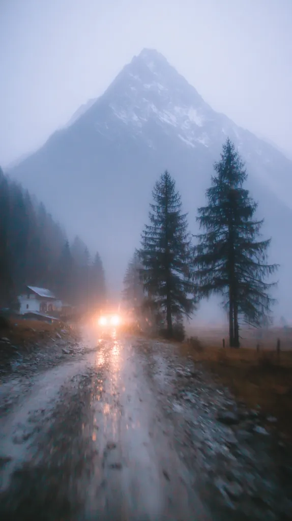 A misty mountain road winds through a valley, leading towards a large, snow-dusted peak.  Two tall evergreen trees stand sentinel beside the wet, muddy path.  Headlights of an approaching vehicle illuminate the road ahead, creating a blurry effect from a moving perspective. The scene is serene yet dramatic, the fog enhancing the mountain's imposing presence. A small building is visible near the roadside.