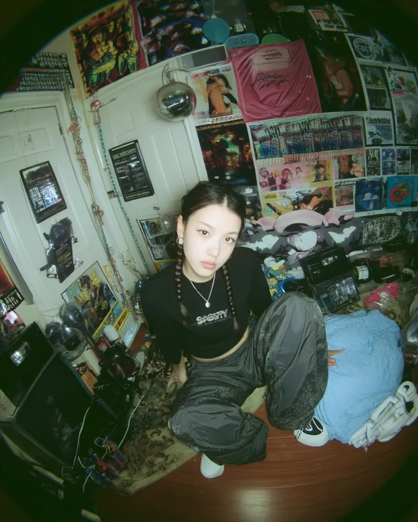 A young woman with braided hair sits in a room cluttered with posters, records, and other memorabilia.  She wears a black long-sleeved crop top, dark cargo pants, and white sneakers. The fisheye lens distorts the perspective, emphasizing the chaotic yet stylish atmosphere of the room.  Her expression is calm and direct, contrasting the vibrant background.