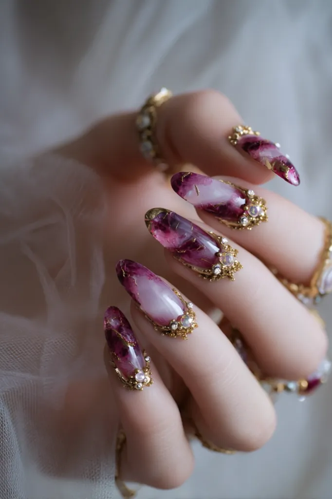 Close-up of elegantly manicured nails.  Each nail features a long, almond shape with a deep purple and white marbled design.  Intricate gold detailing and small, sparkling embellishments adorn the base of each nail, creating a luxurious, jewel-toned effect. The hand is delicately posed against a sheer, light-colored fabric.  The overall aesthetic is romantic and sophisticated.