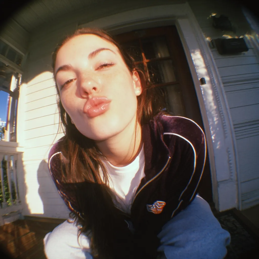 A young woman with long brown hair puckers her lips for a kiss in a close-up, fisheye-lens selfie.  She's wearing a dark purple velvet zip-up jacket with white trim and a white t-shirt underneath.  The background shows a sunlit porch of a house. The image has a vintage, film-like quality.