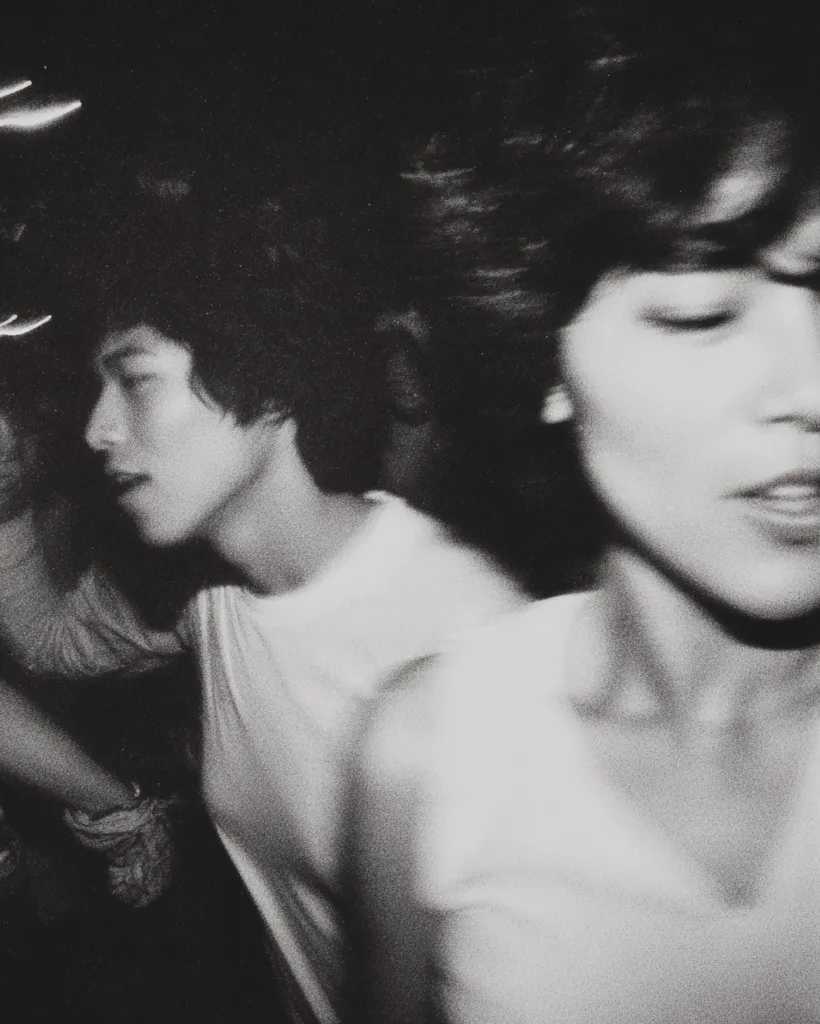 A black and white, blurry photograph captures two young women, possibly in a dimly lit club or concert. The woman in the foreground is slightly out of focus, her face partially obscured, conveying a sense of movement and energy.  The woman in the background is more defined, her profile visible, wearing a simple white t-shirt. The overall impression is one of fleeting moments and youthful vitality.