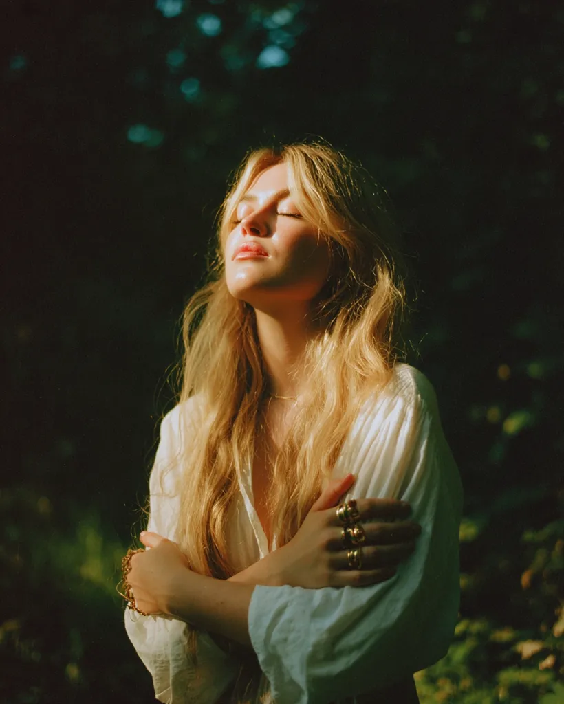 A young woman with long blonde hair stands in a sun-dappled forest.  She wears a loose white shirt and several gold rings. Her eyes are closed, and her arms are crossed over her chest, suggesting serenity and peace.  The warm sunlight illuminates her face, creating a soft, ethereal glow against the dark background. The overall mood is one of quiet contemplation and natural beauty.