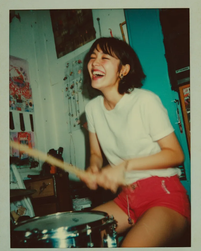A smiling young woman with short dark hair enthusiastically plays the drums in her bedroom.  The room is cluttered with posters and personal items, showcasing a vibrant and youthful aesthetic. She's wearing a white t-shirt and red shorts, and the image has a grainy, nostalgic quality reminiscent of a vintage Polaroid photograph. The overall mood is joyful and energetic.