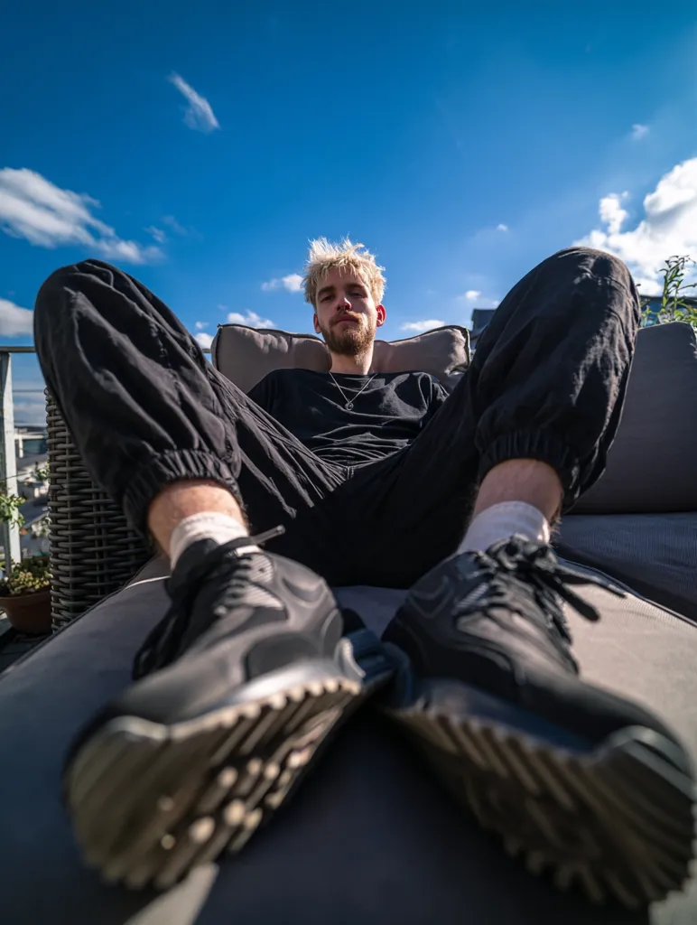 A young man with blond hair relaxes on an outdoor lounge chair against a bright blue sky. He's wearing black joggers and sneakers, a simple black shirt, and a silver necklace. The low-angle shot emphasizes the vastness of the sky and the comfortable repose of the individual. The scene is peaceful and sunny.