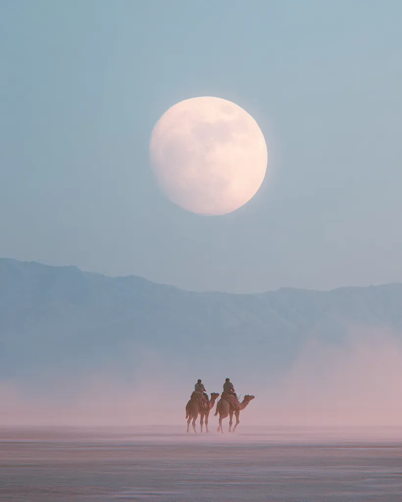 Two figures ride camels across a vast, hazy desert landscape under a large, pale moon.  The scene is serene and peaceful, with muted colors dominating the image.  Mountains are visible in the distance, barely discernible through the atmospheric haze. The overall mood is one of quiet solitude and vastness.