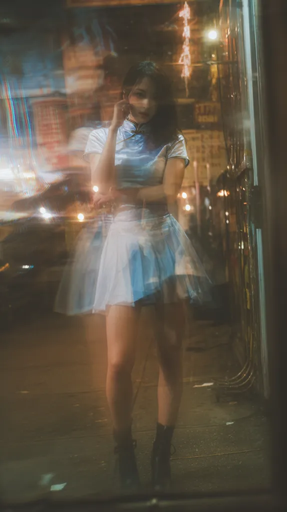 A young woman, viewed through a blurry glass surface, stands on a city street at night. She wears a light blue and white pleated skirt and a matching collared top, evoking a retro aesthetic.  The bokeh effect from city lights creates a dreamy atmosphere. Her expression is pensive, her hand gently touching her face.  The overall impression is one of quiet contemplation in an urban setting.