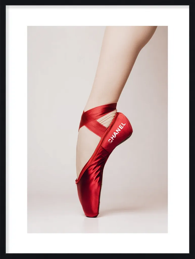 A framed photograph showcases a ballerina's elegantly poised foot, clad in a vibrant red satin pointe shoe.  The shoe is subtly branded with the Chanel logo.  The image is minimalist, with a neutral background highlighting the shoe and the dancer's graceful leg.  The overall aesthetic is chic and sophisticated.