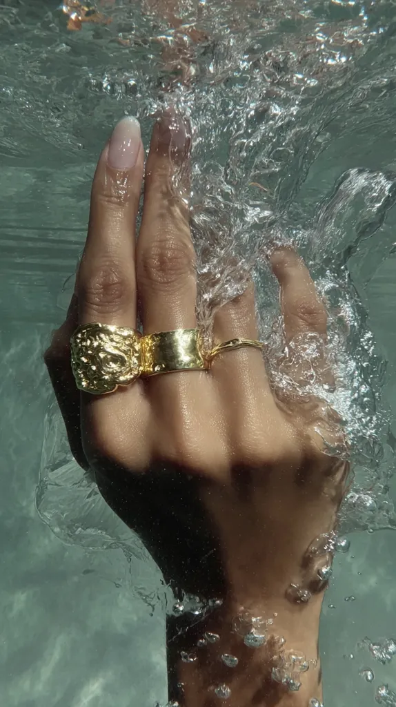 A hand adorned with gold rings emerges from underwater.  The textured gold band is prominent, accompanied by a simpler, thinner ring.  Bubbles cling to the skin and swirl around the hand, creating a dreamy, ethereal effect. The water is a calm, teal-green hue, contrasting beautifully with the gold jewelry.  The overall image evokes a sense of luxury and tranquility.